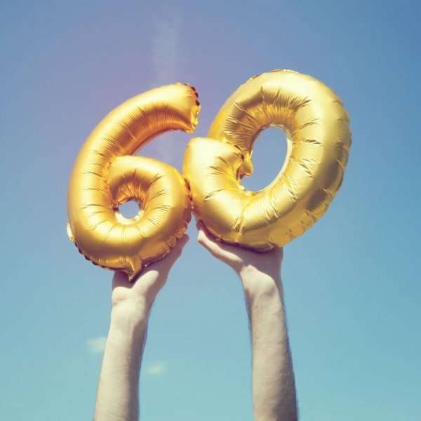 A gold foil number 60 balloon is held high in the air by caucasian male hand.  The image has been taken outdoors on a bright sunny day, the sky is blue with some clouds. A vintage style effects has been added to the image.
